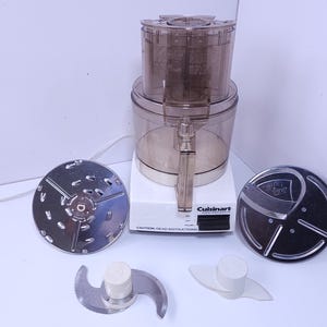 May include: A white Cuisinart food processor with a clear plastic work bowl and lid. Included are two circular metal slicing and shredding discs, a metal S-blade with a white plastic center, and a white plastic dough blade attachment.