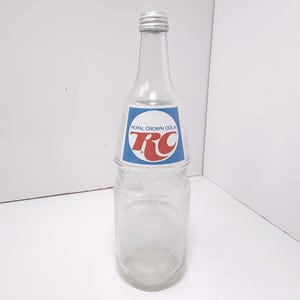 May include: A clear glass Royal Crown Cola bottle with a silver cap. The bottle has a blue and white label with the RC logo in red. The bottle is empty and appears to be vintage.