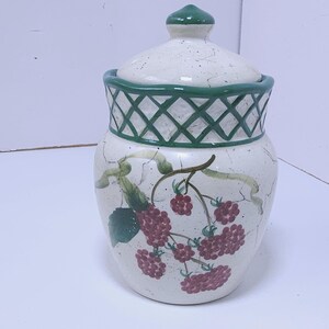 May include: A ceramic jar with a lid, featuring a white base with a green lattice design around the rim. The jar is decorated with a branch of red raspberries and green leaves. The lid has a green knob.