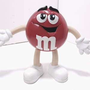 May include: A large, maroon M&M's character figurine with a smiling face, white gloves, and shoes. The letter "m" is prominently displayed on the front. The figurine has tan arms and legs, and black accents on the face.