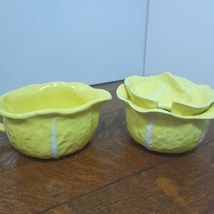 May include: A set of two yellow ceramic creamer and sugar bowls shaped like a head of cabbage. The bowls have white lines on them.