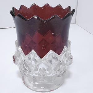 May include: A decorative glass vase with a deep red upper section and a clear, diamond-patterned base. The rim has a scalloped edge. The vase is designed to hold flowers or other decorative items. The vase is made of glass.