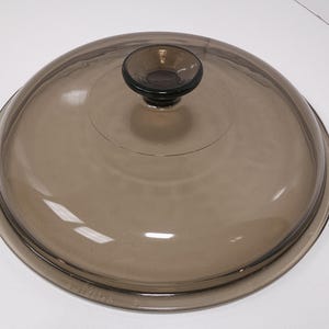 May include: A round, brown glass lid with a small, dark knob. The lid is slightly domed and has a dark rim. The glass is transparent, allowing a view of the contents below. A kitchenware item.
