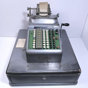 May include: An antique silver-toned mechanical calculator with a paper roll and numerous black and green buttons. The calculator has a drawer with a keyhole and a black handle on the side. A small paper label is attached to the front.