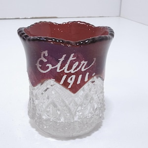 May include: A vintage glass tumbler with a clear, cut-glass base and a deep burgundy rim. The words "Etter 1911" are etched in white script on the upper portion of the glass. The glass has a scalloped rim.