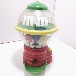 May include: A colorful M&M's candy dispenser. The dispenser features a clear globe with the M&M's logo, a yellow ring, and a red base. Inside the globe are two M&M's characters. The dispenser has a green top and a green tray at the bottom.