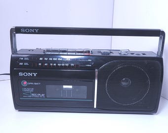 Może przedstawiać: Czarny boombox Sony ze srebrnym uchwytem, głośnikiem i panelem sterowania. Radio posiada tuner AM/FM i odtwarzacz kaset. Logo Sony jest widoczne z przodu i na górze.