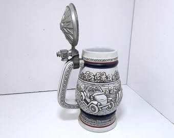 May include: A ceramic beer stein with a hinged metal lid. The stein features a black and white illustration of a vintage car and the text "1913 Stanley Steamer 1913". The handle is white with a black pattern.