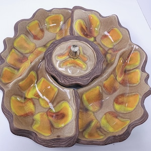 May include: A ceramic divided serving platter with a central lidded bowl featuring a metallic knob. The dish has a wavy, organic edge and a speckled tan glaze decorated with abstract yellow and orange petal-like patterns across the surface.