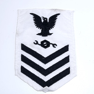 May include: White patch with black embroidered details. Features a black eagle at the top, a wrench and gear symbol, and three chevrons. This patch is likely a military insignia, possibly for a specific rank or trade.