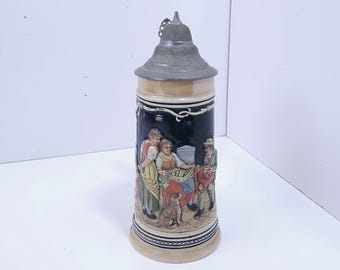 May include: A tall ceramic beer stein featuring a dark blue body with a relief scene of three figures and a dog. The stein is topped with a pointed, embossed metal lid and has a light beige base and rim with decorative black line patterns.