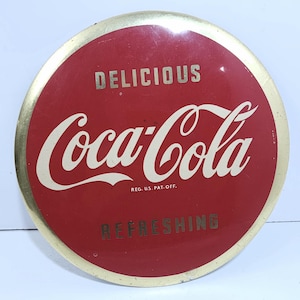 May include: A round, red and gold Coca-Cola sign with the text "Delicious Coca-Cola Refreshing" and "Reg. U.S. Pat. Off."