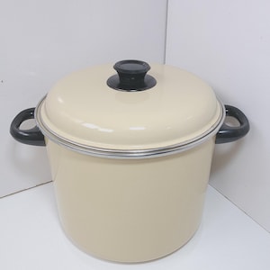 May include: A cream-colored enamel pot with a matching lid and black handle grips. The pot has a silver rim and a black knob on the lid. This cooking pot is suitable for various kitchen tasks.