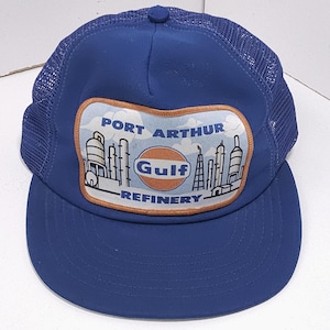 May include: A blue trucker hat with a mesh back and a curved brim. The front features a patch with the text "PORT ARTHUR GULF REFINERY" and a logo. The hat is a classic style, suitable for casual wear.