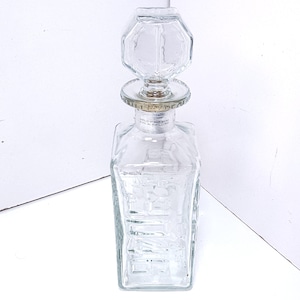 May include: A clear glass decanter with a geometric stopper. The decanter has a rectangular body with etched details and a silver-colored band around the neck. The stopper is a faceted, octagonal shape. The decanter is empty.