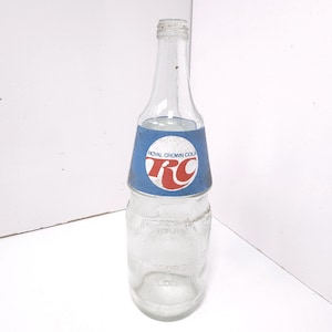 May include: An empty, clear glass Royal Crown Cola bottle. The bottle has a blue and white label with the words "Royal Crown Cola" and the letters "RC" in red. The bottle is empty and has a vintage appearance.