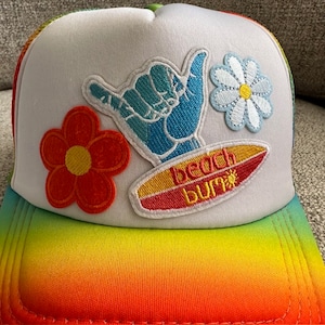 May include: White trucker hat with a rainbow ombre brim. The hat features embroidered patches of a flower, a hand giving the shaka sign, and a surfboard with the text "beach bum".