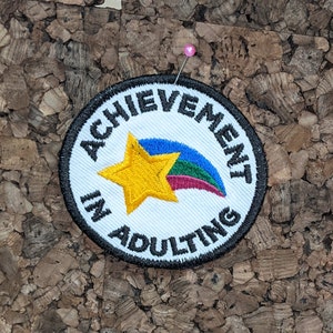 May include: A white embroidered patch with black text that reads "Achievement in Adulting". The patch features a yellow star with a rainbow-colored comet tail.