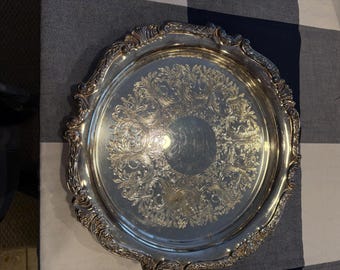 May include: A silver serving tray with an ornate, embossed design. The tray has a raised rim and a scalloped edge.