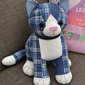 May include: A stuffed cat toy crafted from blue plaid and solid white fabric. The cat features a white face patch, a pink stitched nose, black button eyes, and a black collar with white dots. The paws and tail tip are white with stitched details.