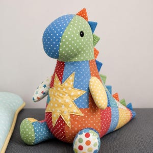 May include: A stuffed dinosaur toy constructed from various colorful fabric patches featuring star, dot, and cross patterns. The creature has a green head, a yellow star appliqué on its chest, and a row of triangular spikes along its back.