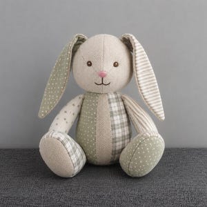 May include: A stuffed rabbit toy made from various fabric patches in shades of beige, sage green, and white. The rabbit features a round head, long ears with polka dot and striped patterns, and a body constructed from herringbone, plaid, and star-print fabric.