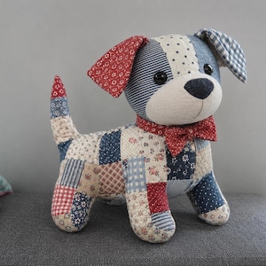 May include: A stuffed dog toy crafted from various patchwork fabric squares in shades of red, blue, and cream. The dog features a red bowtie, black button eyes, a black nose, and ears with contrasting patterns including stars and gingham checks.