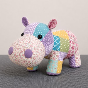 May include: A stuffed hippopotamus toy constructed from various fabric patches in floral, gingham, polka dot, and geometric patterns. The colors include shades of purple, teal, yellow, and pink. It features two black button eyes and purple felt nostrils.