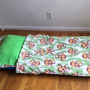 May include: A children's nap mat set featuring a green pillow and a blanket with a white background. The blanket is decorated with brown monkeys and green leaves. The set is designed for comfort and convenience.