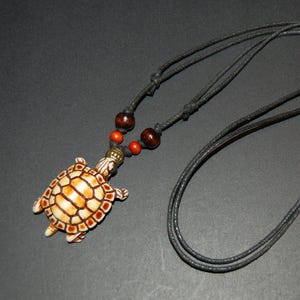 Puede incluir: Un collar con colgante de tortuga y cordón negro. El colgante de tortuga es de color crema con detalles marrones y un diseño de caparazón detallado. El collar también presenta cuentas de madera en marrón y naranja.