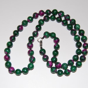 May include: A beaded necklace featuring round, polished stones with a mottled pattern of deep forest green and vibrant magenta. The beads are strung on a dark cord with individual knots between each stone, secured by a silver-toned metal clasp.