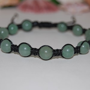 Puede incluir: Un brazalete de cuentas de piedra verde con cordón negro. El brazalete está anudado con un estilo macramé.