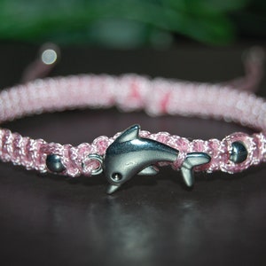 Puede incluir: Un brazalete trenzado rosa con un dije de delfín plateado.