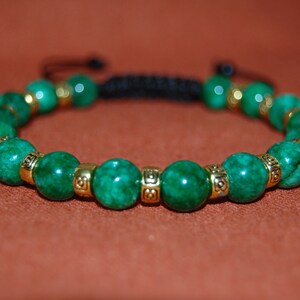 Puede incluir: Un brazalete de piedras preciosas verdes con detalles dorados y cierre de cordón negro.