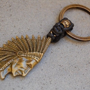 May include: A gold-toned keychain featuring a detailed profile of a Native American wearing a feathered headdress. The keychain is attached to a black braided cord with a gold bead.