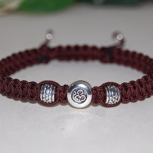 May include: A maroon braided bracelet with silver-colored accents. The bracelet features a central silver charm with an Om symbol and two decorative silver beads. The adjustable cord bracelet is a stylish accessory.