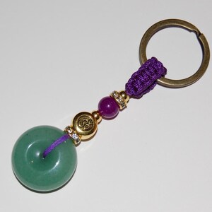 Puede incluir: Un llavero de piedra de jade verde con un cordón morado y un dije dorado. El dije tiene un símbolo y está rodeado de pequeños diamantes de imitación.