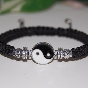 Könnte beinhalten: Schwarzes geflochtenes Armband mit einem silbernen Yin-Yang-Anhänger und zwei silbernen Perlen mit einem Wirbelmuster.