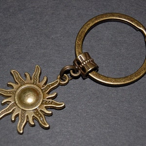 Könnte beinhalten: Ein bronzefarbener Schlüsselanhänger mit einem Sonnenmotiv. Die Sonne hat eine erhabene Mitte und Strahlen, die sich nach außen erstrecken. Der Schlüsselanhänger ist an einem Metallring befestigt.