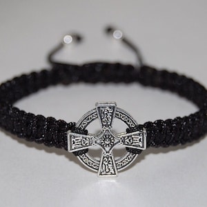 May include: A black braided bracelet with a silver Celtic cross charm. The cross is detailed with intricate designs and has a circular border.