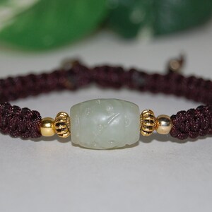 Puede incluir: Un brazalete trenzado marrón oscuro con una cuenta de jade verde claro y detalles dorados.