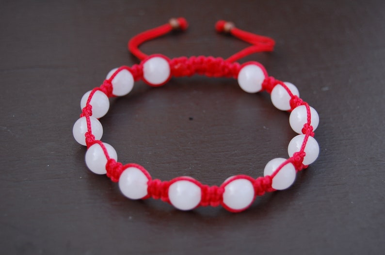 May include: A red string bracelet with white beads. The bracelet is tied with a knot and has a tassel on the end.
