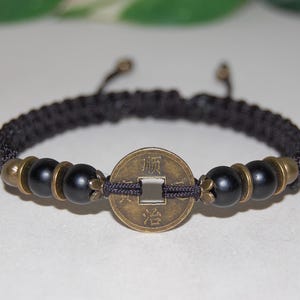 May include: Black braided bracelet with a central bronze coin and black beads. The coin has Chinese characters. The bracelet has bronze-colored accents and an adjustable closure. A decorative accessory.