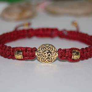 Puede incluir: Un brazalete trenzado rojo con un dije de oro con el símbolo Om y dos cuentas doradas.