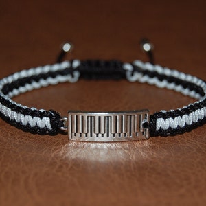 以下が含まれることがあります： 黒と白の編み込みブレスレットに、銀のピアノ鍵盤のチャームが付いている。