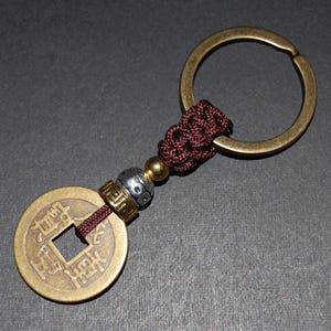 May include: A brass Chinese lucky coin keychain with a square hole in the centre and Chinese characters. The coin is attached to a brown cord and a brass key ring.