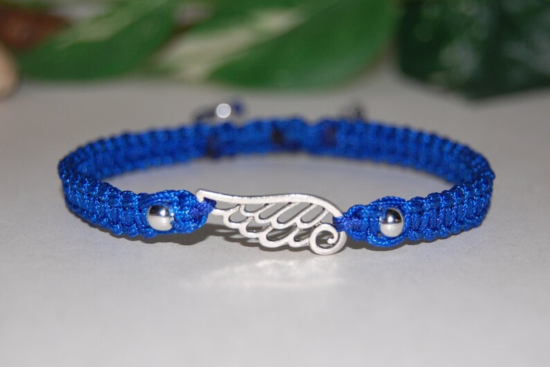 May include: A blue macrame bracelet with a silver wing charm. The bracelet features a woven design and small silver beads at each end of the charm. The wing charm is a detailed, openwork design.