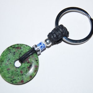 May include: A green and black stone keychain with a silver ring and black cord. The stone has a hole in the center and is surrounded by a blue and white ceramic bead.