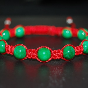 Puede incluir: Un brazalete de cuerda roja con cuentas de piedras preciosas verdes. El brazalete está anudado con un estilo macramé.