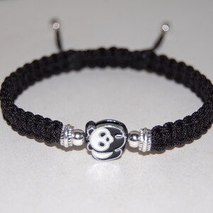 Puede incluir: Pulsera trenzada negra con un dije de plata con dise&ntilde;o de oso panda.